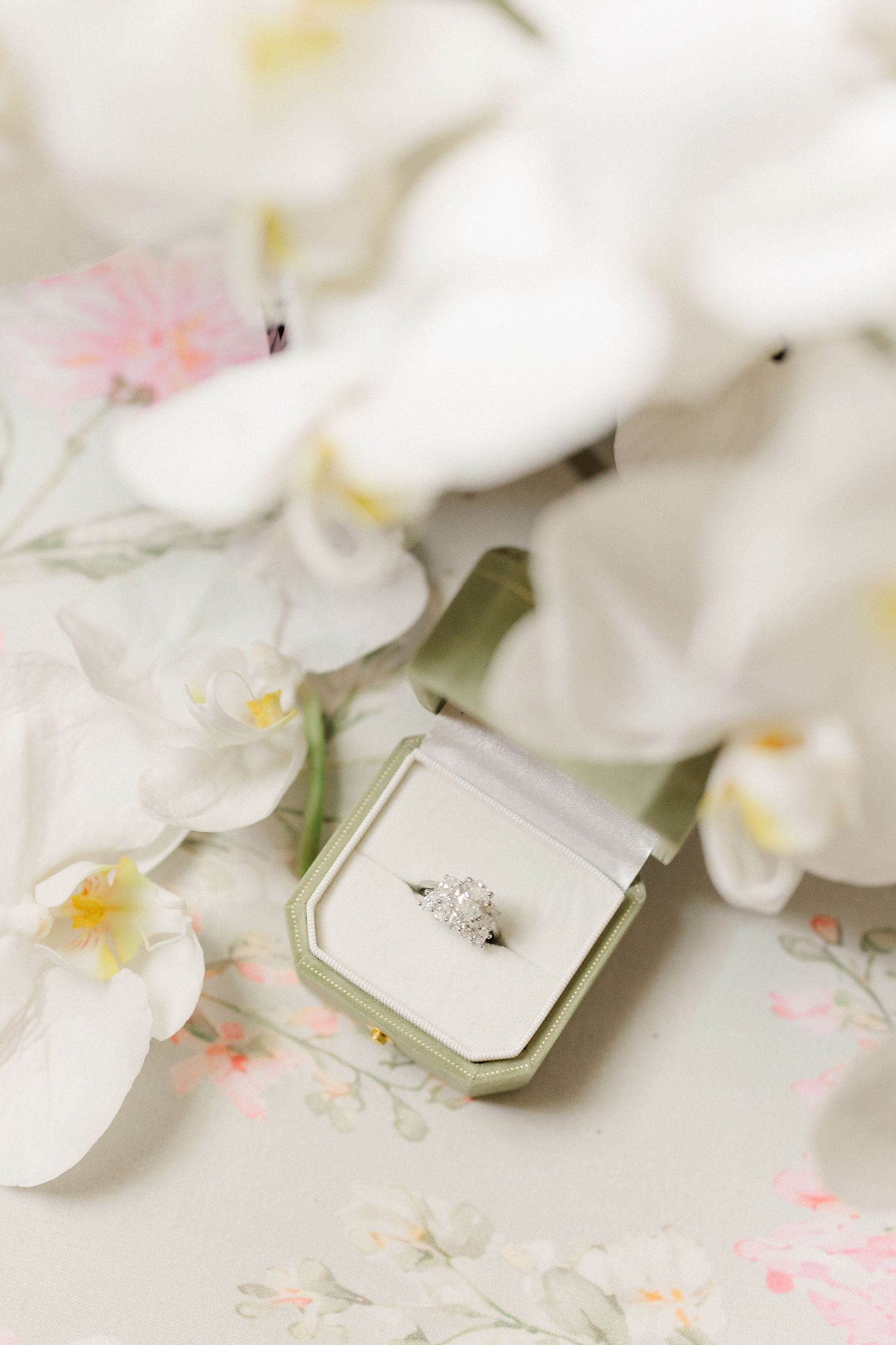 Diamond engagement ring in sage green velvet box surrounded by white orchids on floral surface.