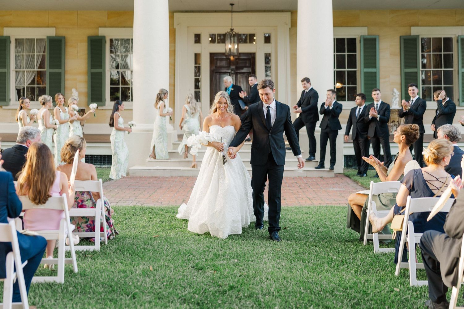 Bride and groom walk back down the aisle as guests applaud at outdoor wedding ceremony on lush green lawn.