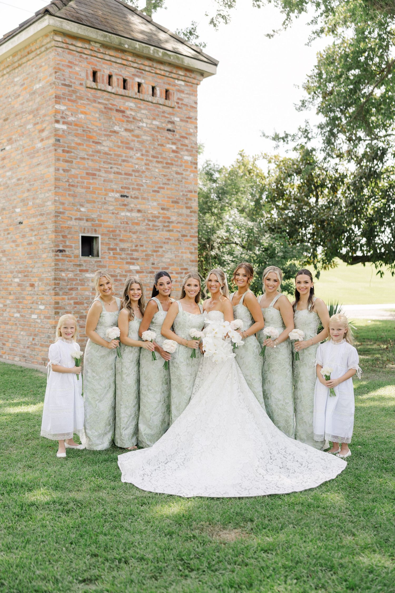 Bride in lace ballgown poses with bridesmaids in sage green dresses and flower girls before brick tower outdoors.