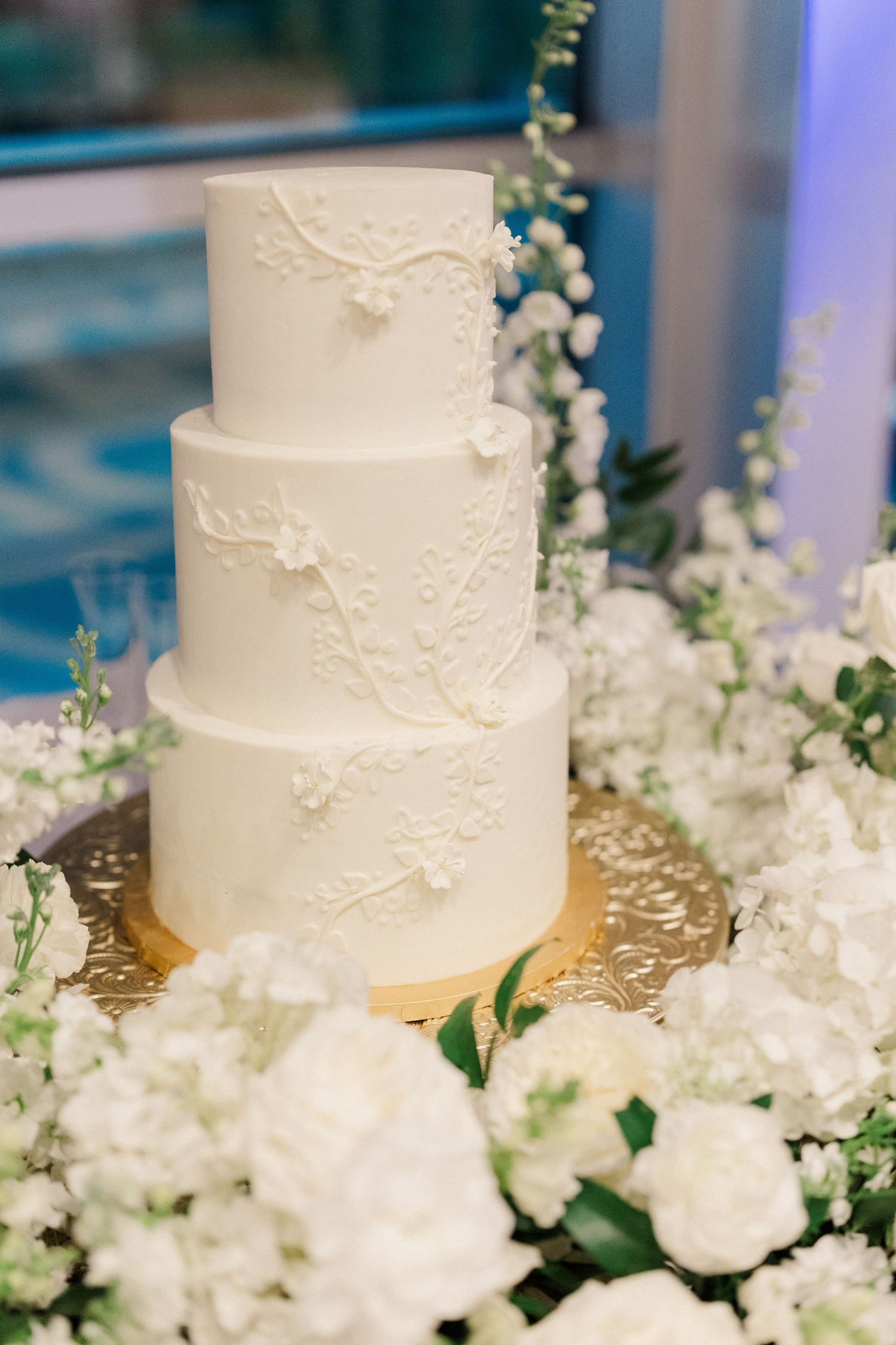 Elegant four-tier white wedding cake with floral appliques surrounded by white roses and baby's breath on a gold stand.