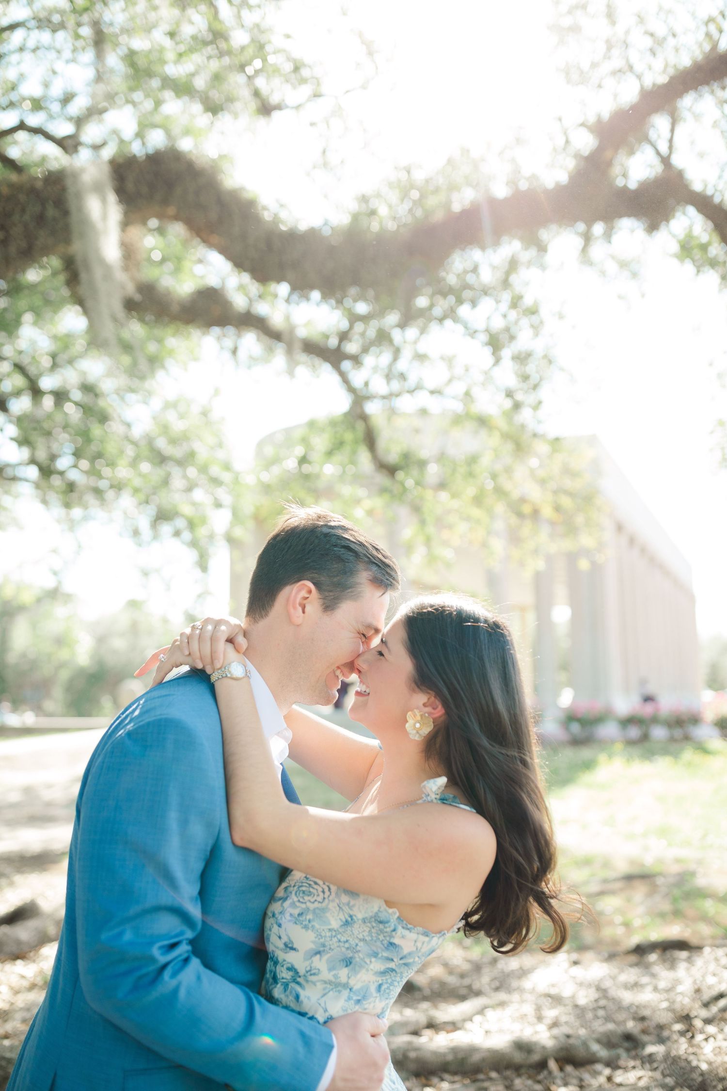 A romantic couple embraces under the dappled sunlight of a large tree branch on a bright sunny day.