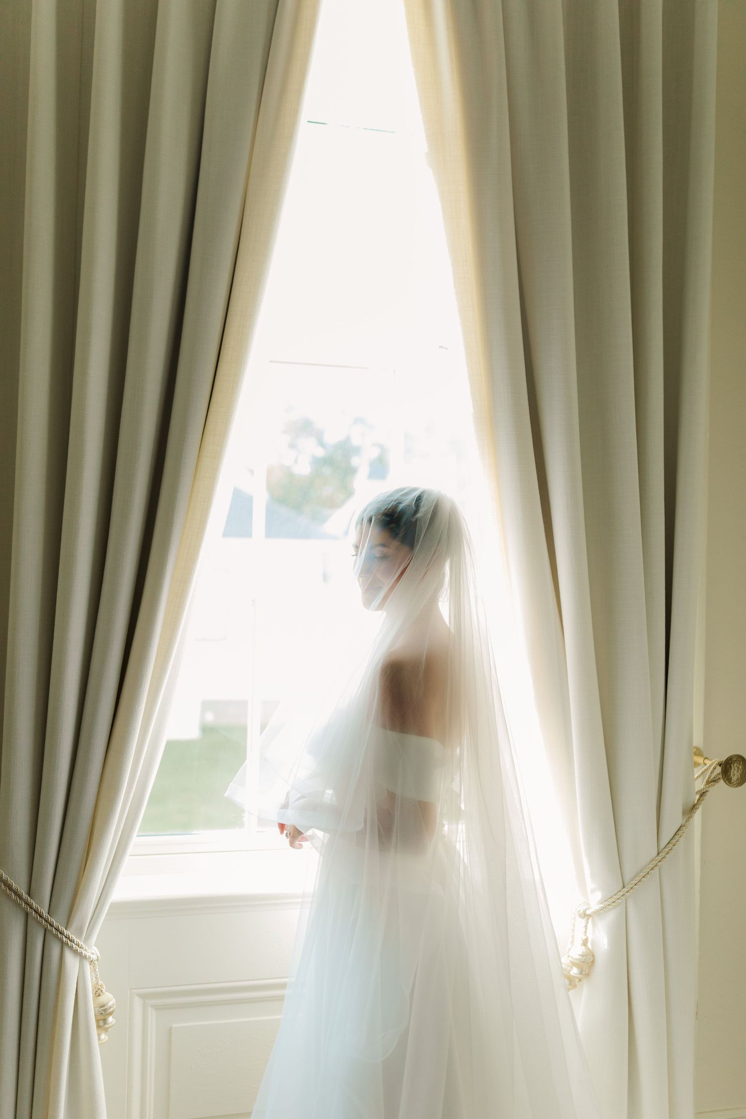 Ethereal silhouette in wedding dress and veil stands by sunlit window with elegant cream curtains.