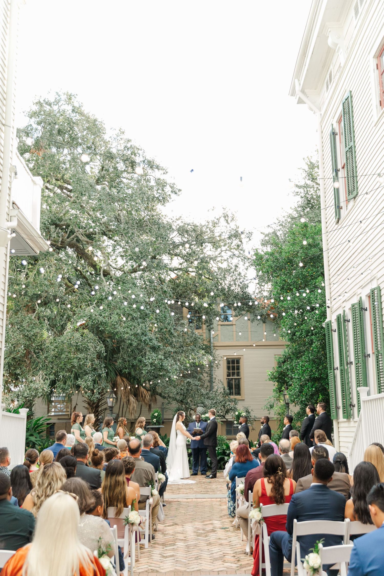 An outdoor wedding ceremony in a courtyard with string lights overhead and brick paving surrounded by historic buildings.
