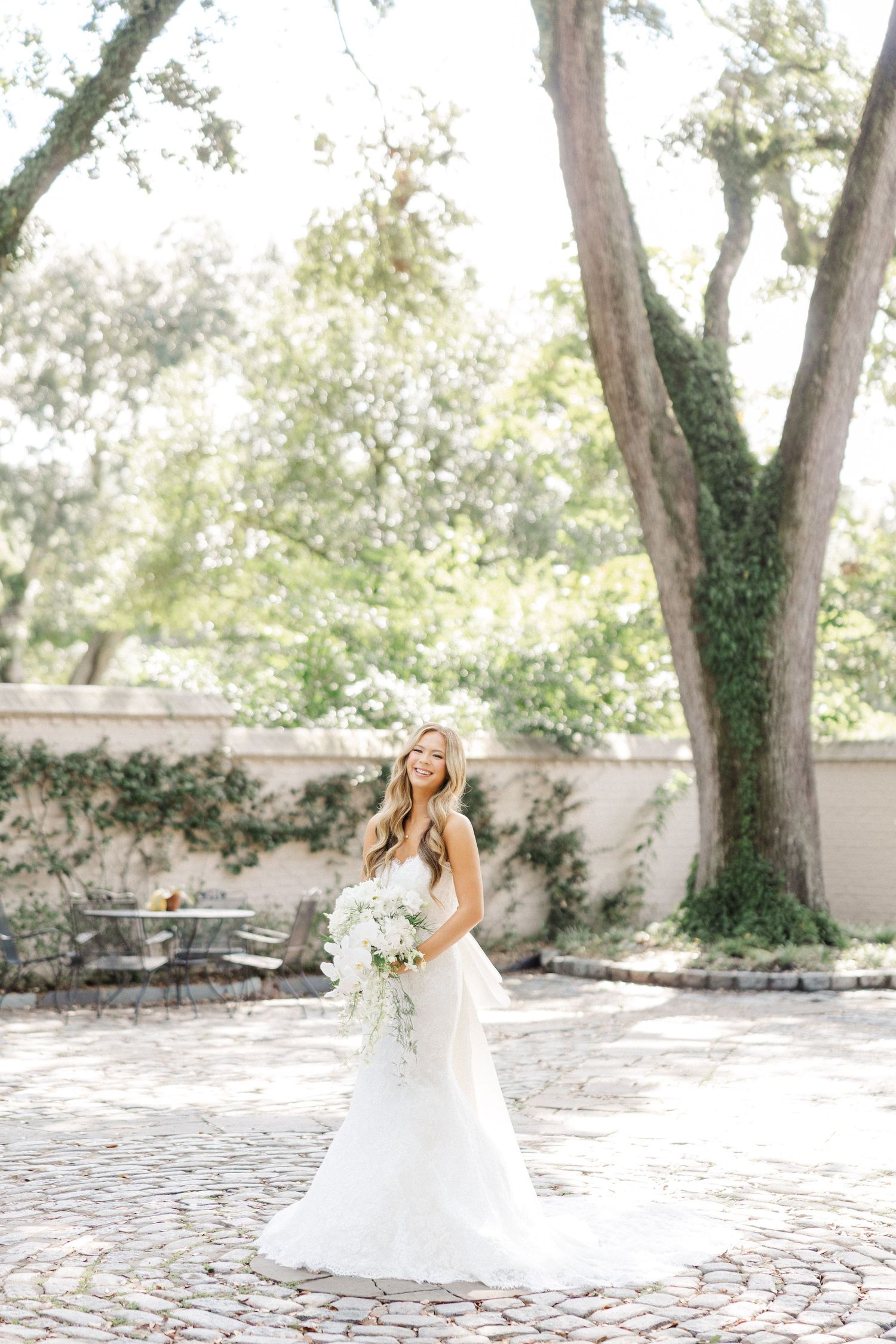 A bride in a flowing white wedding dress poses in a garden courtyard with stone pavers and tall trees.