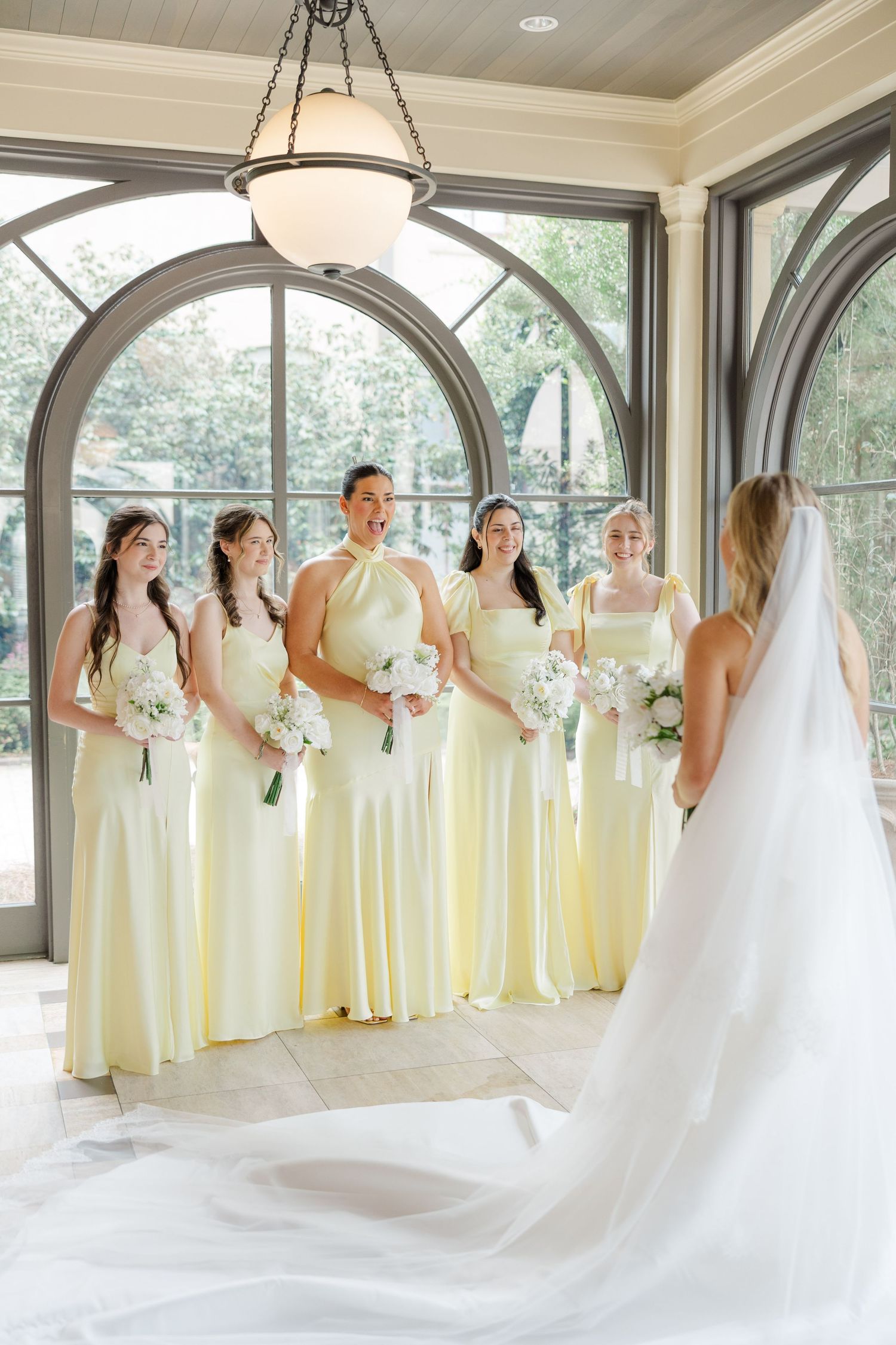 Bridesmaids in pale yellow dresses admiring a white wedding gown in an elegant room with arched windows.
