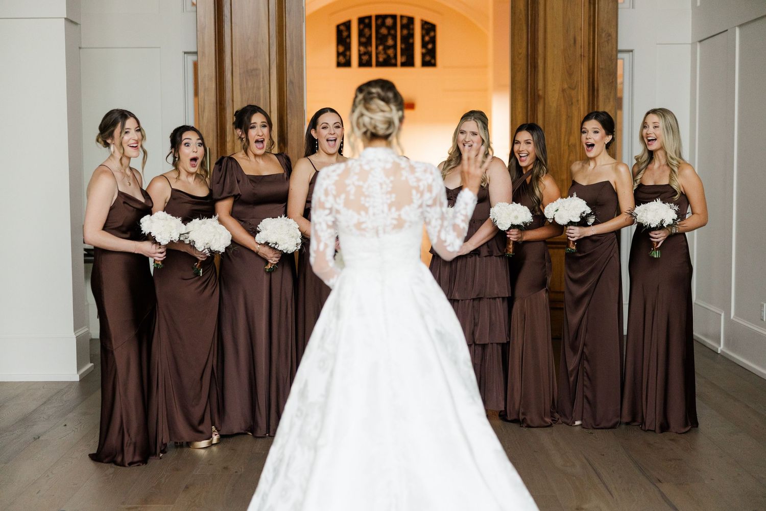 Bridesmaids in chocolate brown dresses line up along a bright hallway as the bride's white gown trails behind.