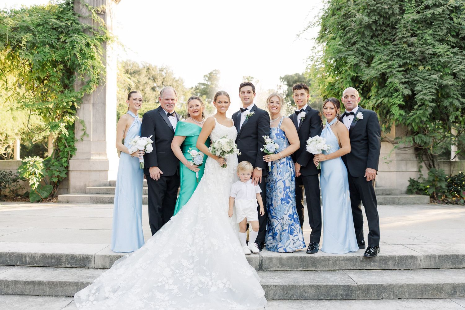 Wedding party in light blue and black formal attire gathers for outdoor group photos.