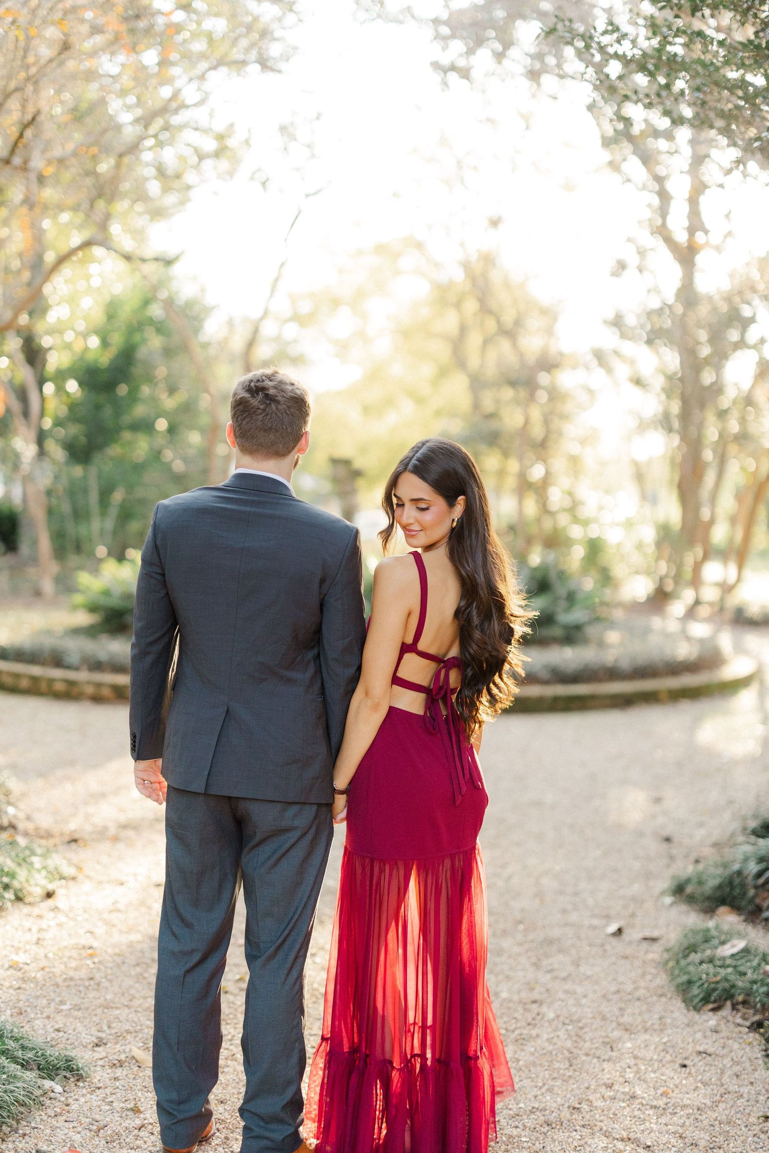 Couple stands on garden path with her burgundy gown featuring strappy back detail against natural backdrop.