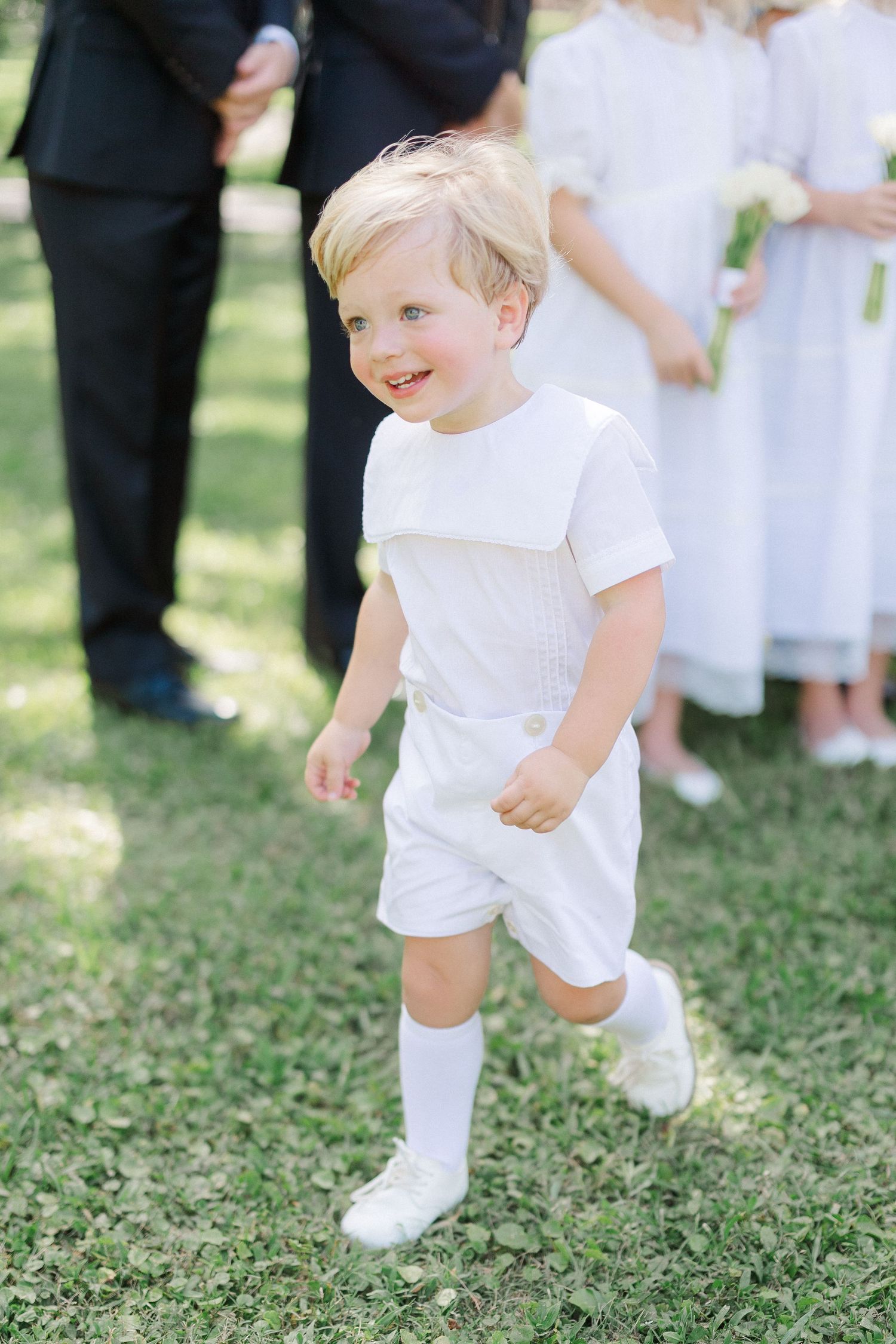 Adorable blond ring bearer in white outfit and knee socks running on green grass at outdoor wedding.