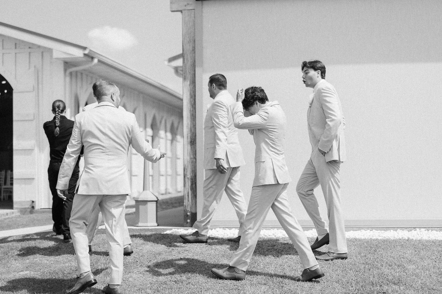 Black and white photo of groomsmen in white suits laughing and joking outside wedding venue.