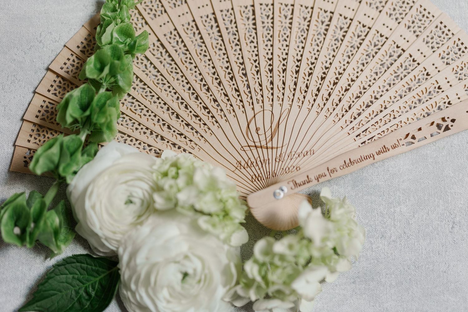 Elegant wooden laser-cut hand fan with white ranunculus and green flowers, wedding favor gift on marble surface.