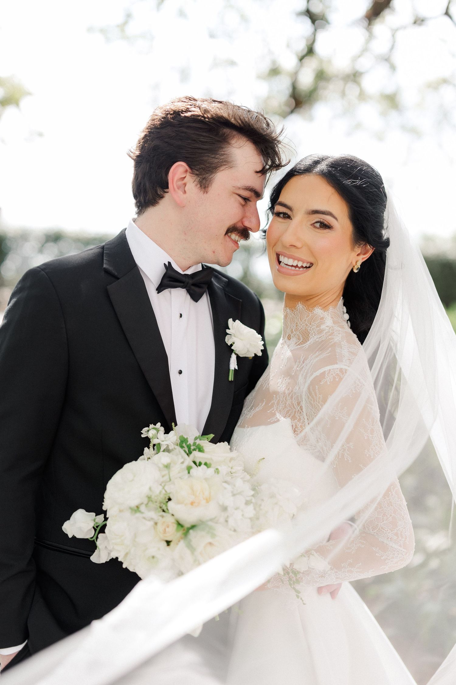 Bride and groom sharing romantic moment, bride holding white bouquet with veil flowing outdoors.