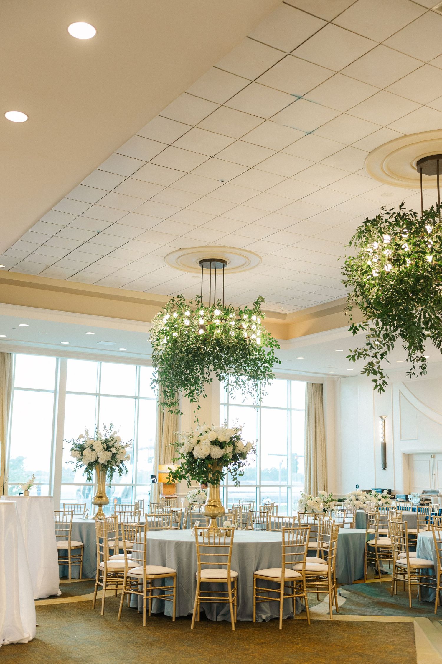 Elegant wedding reception hall with round tables, gold chiavari chairs, lush hanging greenery chandeliers, and soft blue linens.
