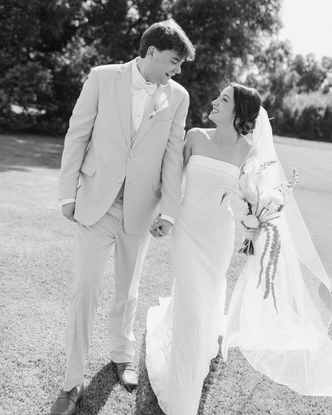 Black and white photo of a smiling bride and groom holding hands on their wedding day outdoors.