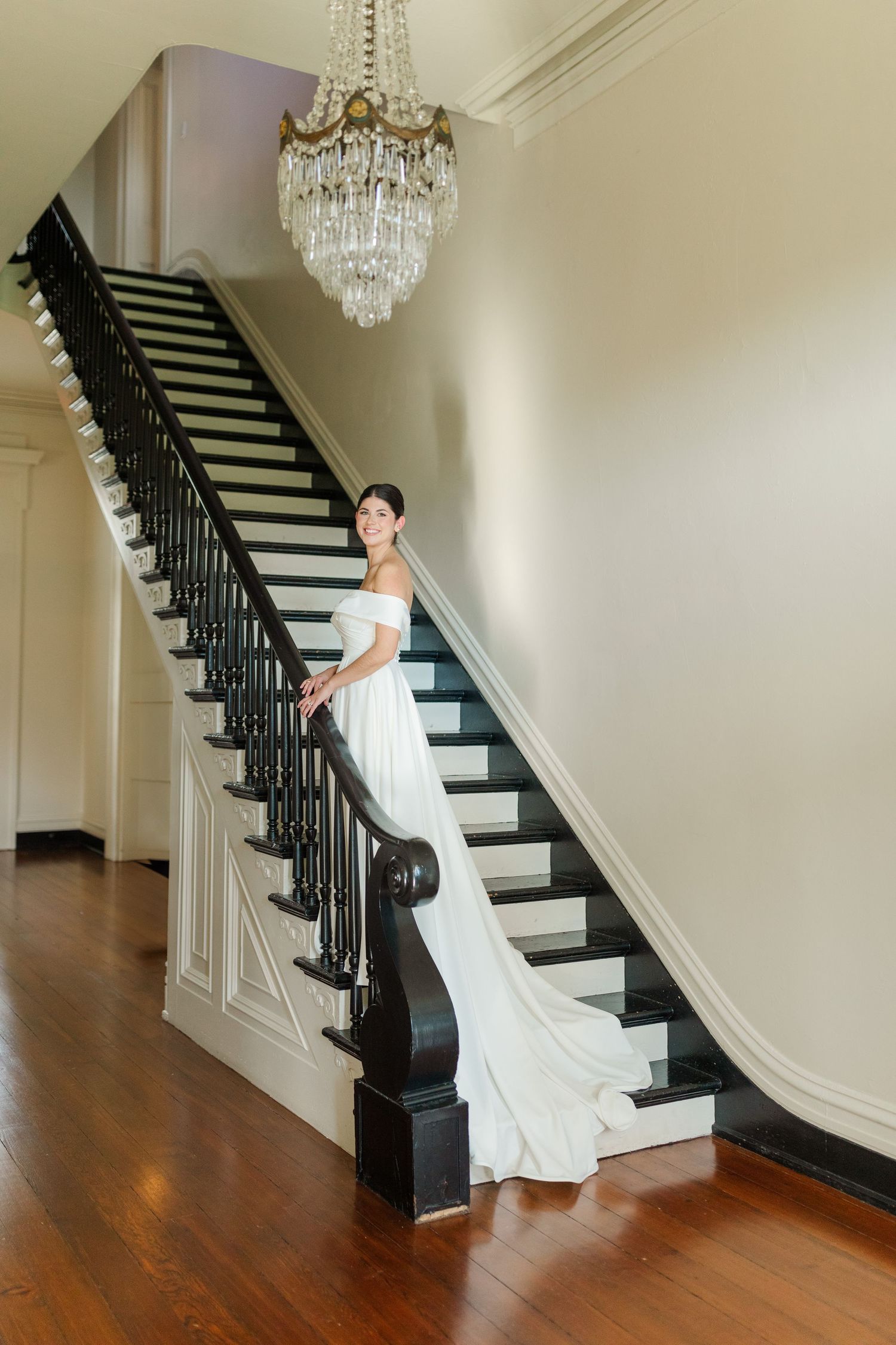 Elegant bride in white gown poses on grand staircase with crystal chandelier in classic indoor setting.
