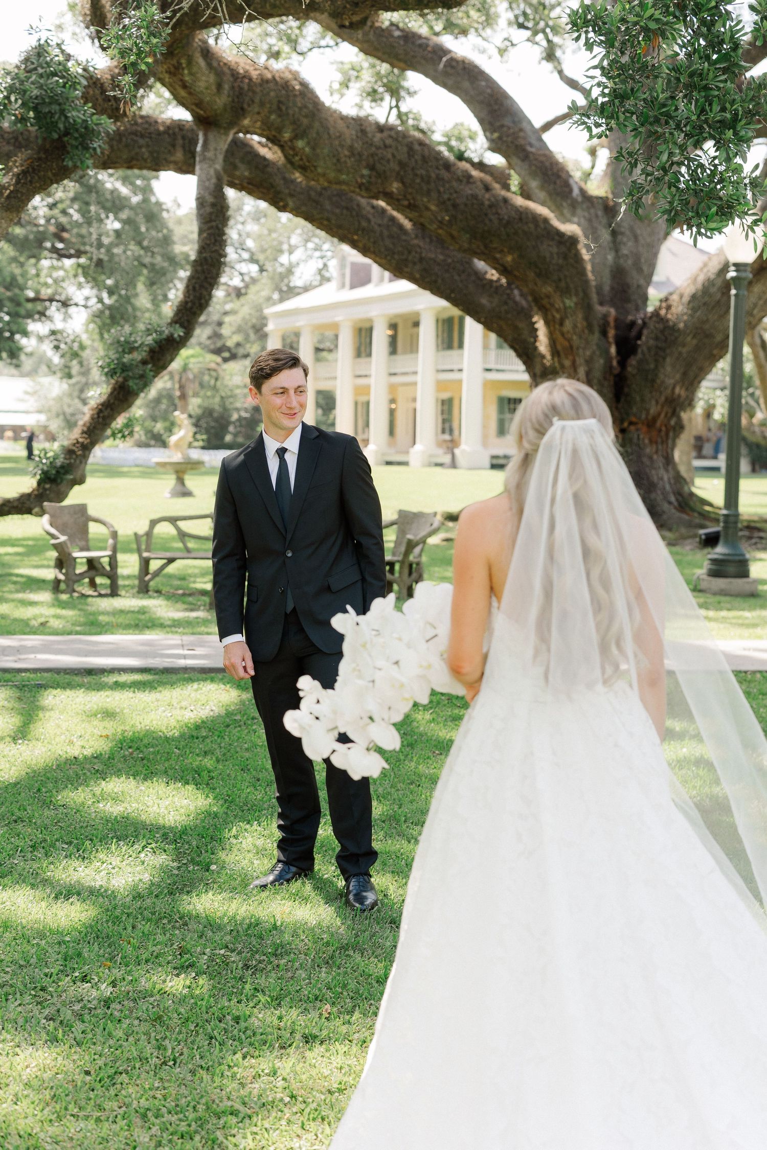 Groom in black suit smiles at bride in white gown and veil holding orchids under oak tree.