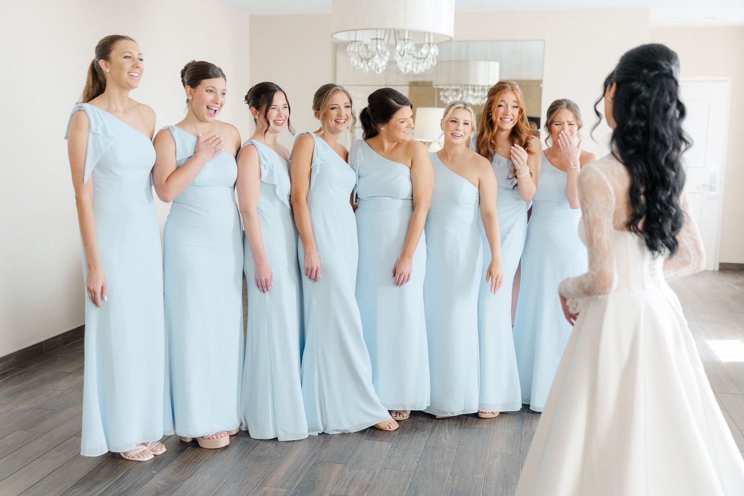 Bride in white gown faces smiling bridesmaids in light blue dresses during wedding morning reveal in bright room.