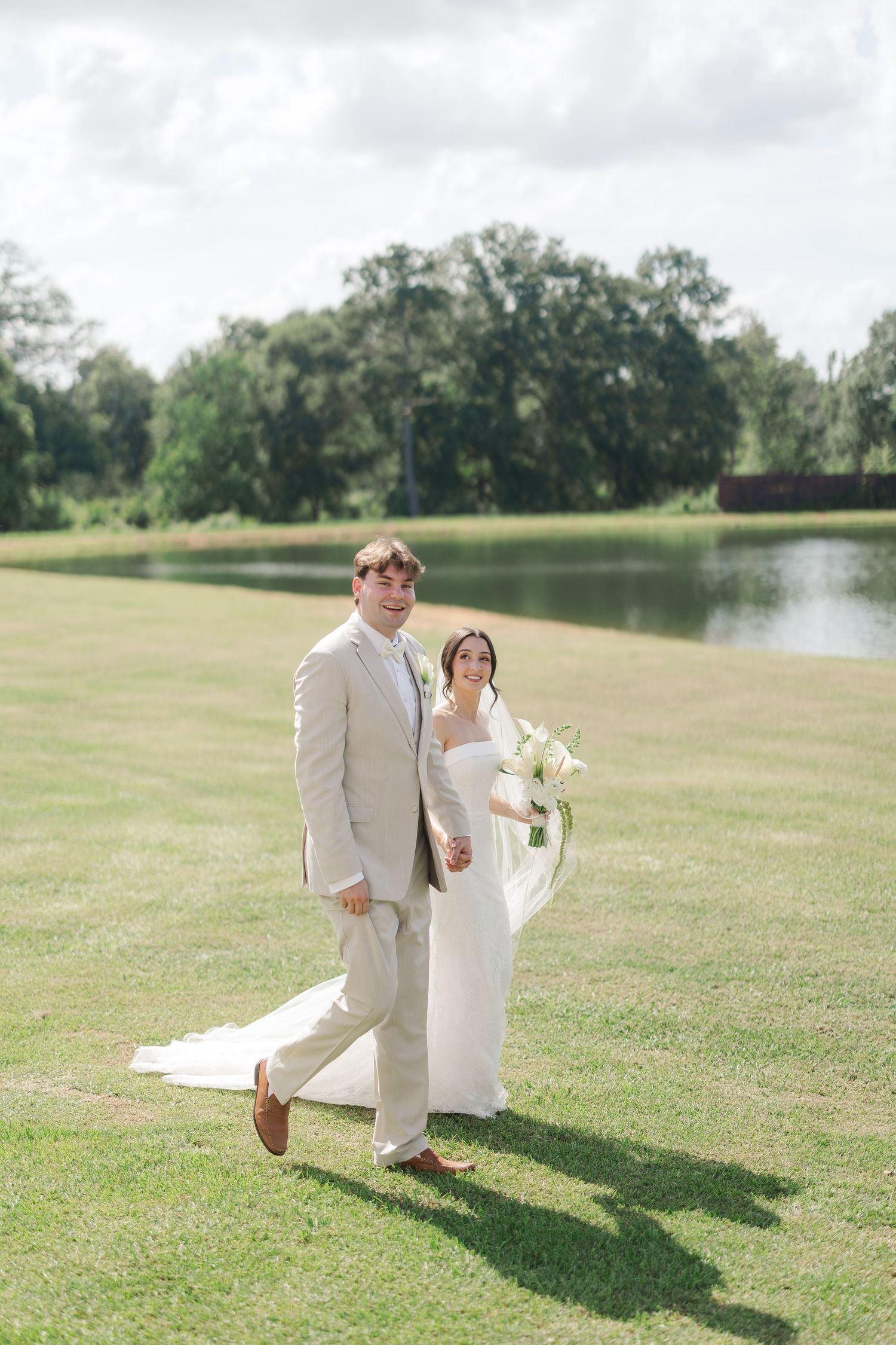 Bride and groom walking on green lawn near pond, bride holding white bouquet with flowing veil.