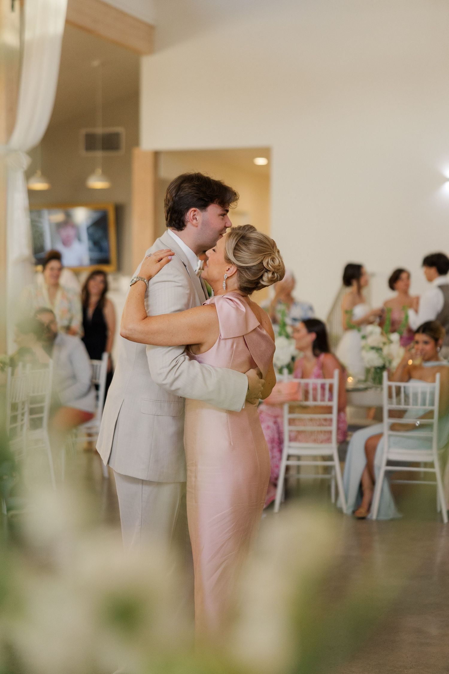 Couple shares romantic kiss during first dance at wedding reception with guests seated at decorated tables.