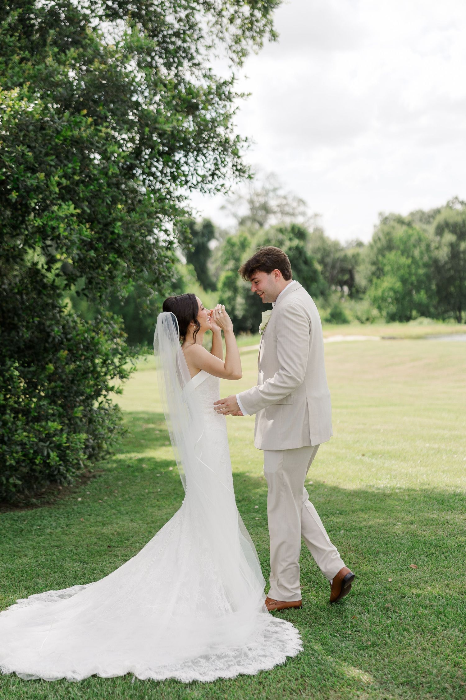 Bride and groom share romantic first look moment on lush green lawn, bride in lace-trimmed cathedral veil.