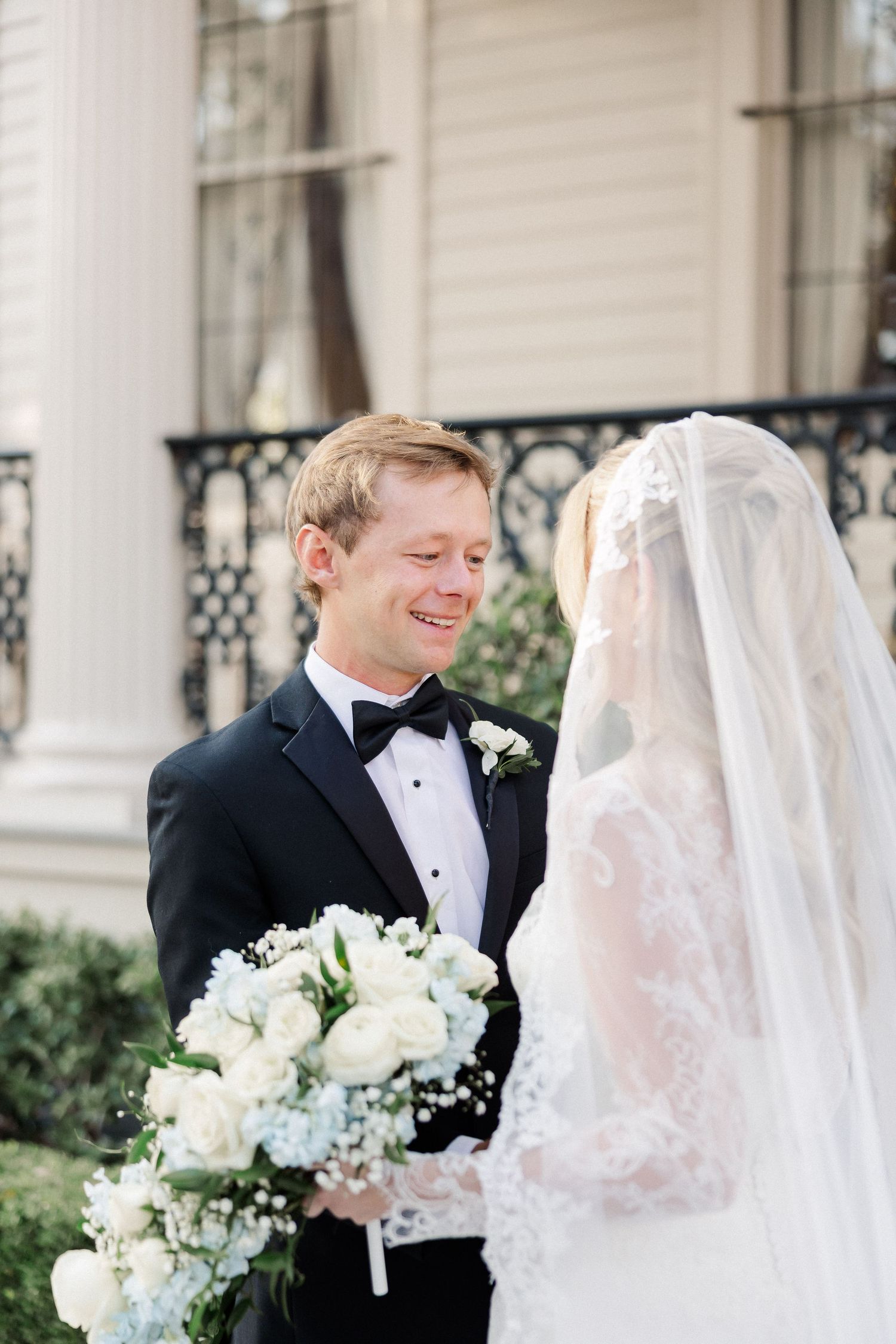 A romantic wedding first look moment with white floral bouquet and elegant architecture in the background.