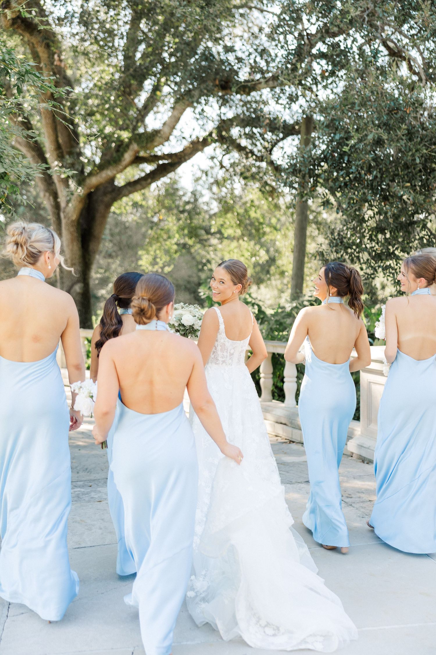 Bridesmaids in flowing light blue dresses walk arm-in-arm down a sunlit garden path.