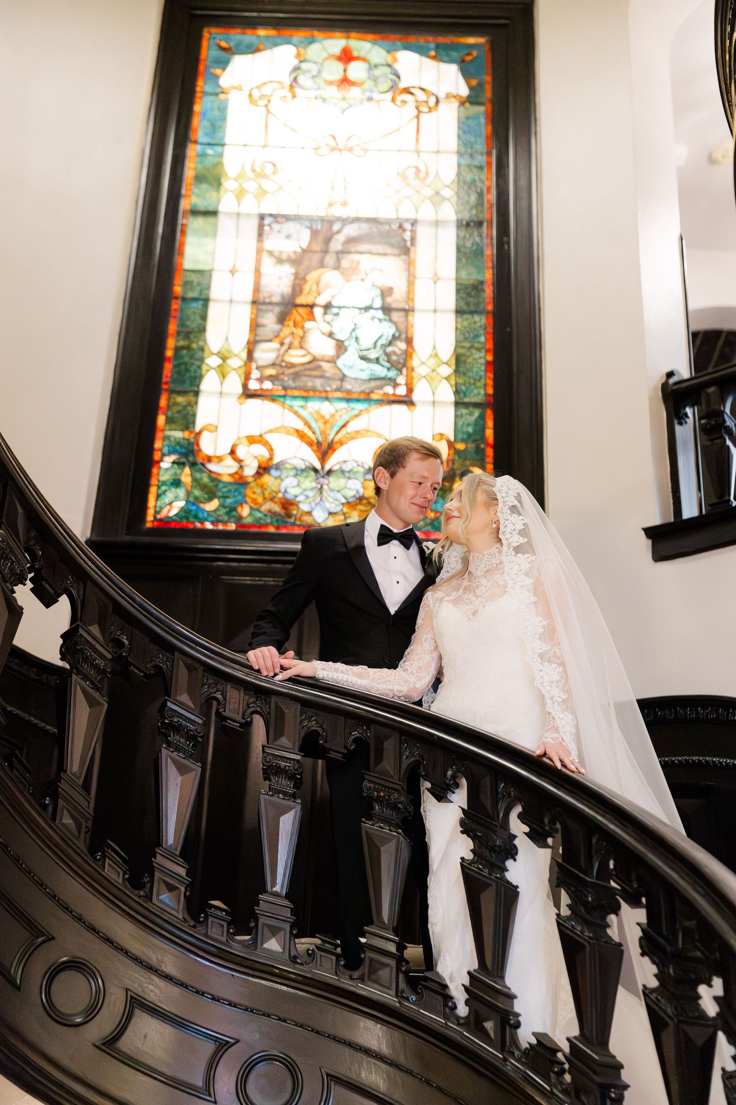 Couple poses on an elegant black staircase with stained glass windows in the background.