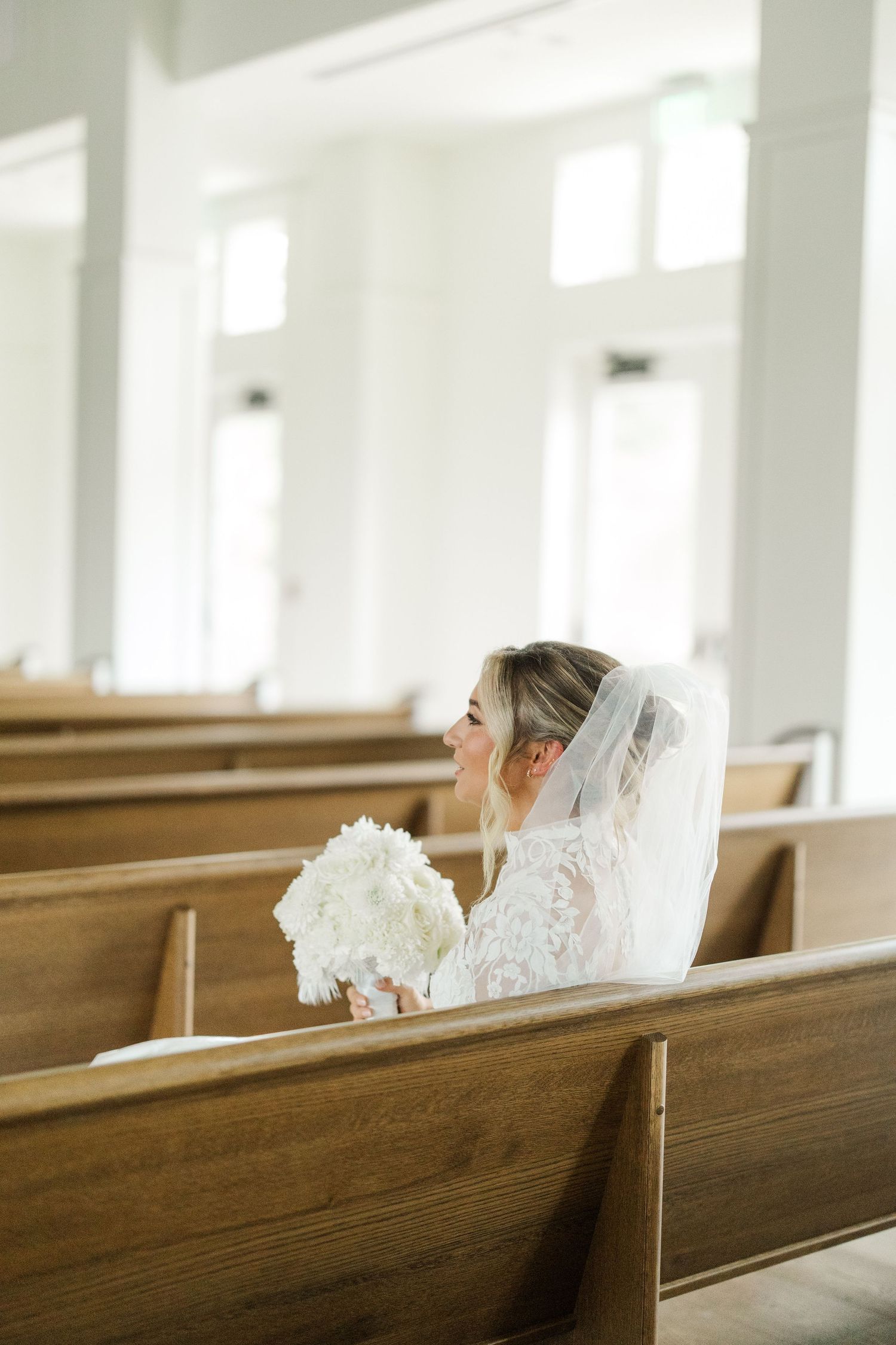 A bride in a lace dress and veil sits in a church pew holding a white bouquet.