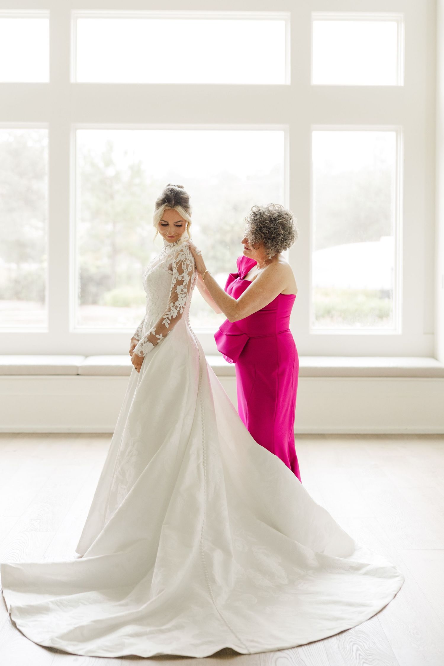 A wedding dress preparation scene with a bright pink-clad helper adjusting the long train in a sunlit room.