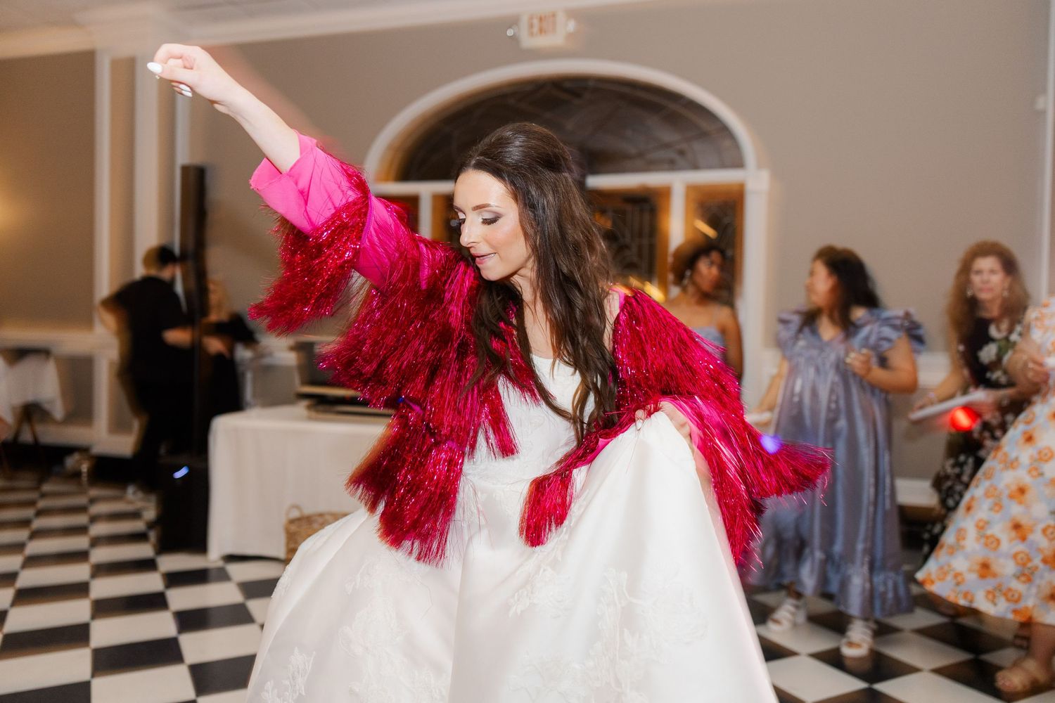 A bride wearing a red fringe jacket dances enthusiastically on a black and white checkered dance floor.