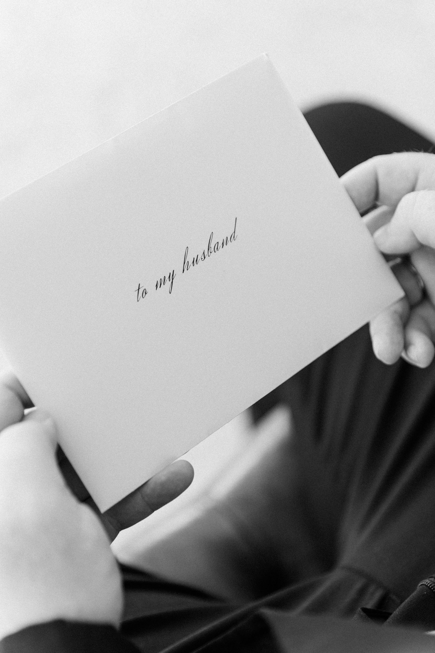 A close-up black and white photo of hands holding a note card with 'to my husband' written in cursive script.