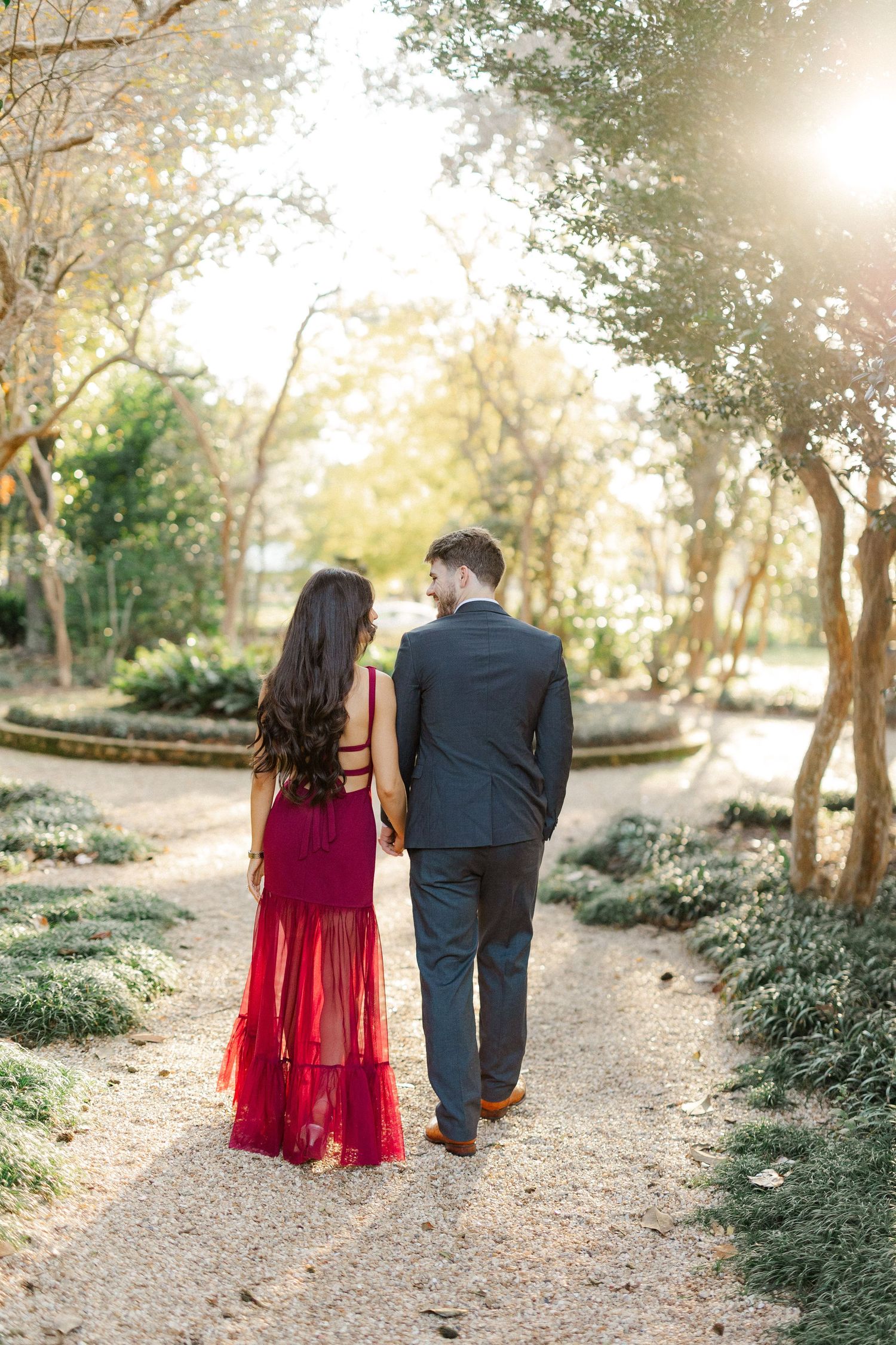A romantic couple strolls down a sunlit garden path, with her in a burgundy gown and him in a dark suit.