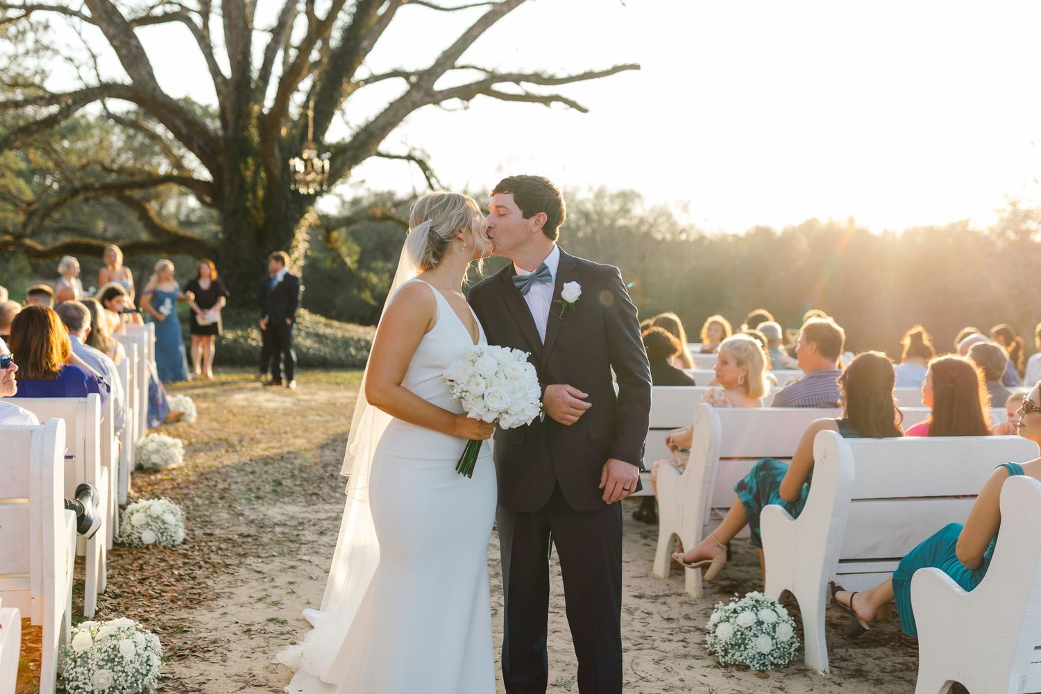 Romantic outdoor wedding ceremony at sunset with guests seated in white chairs under a large oak tree.