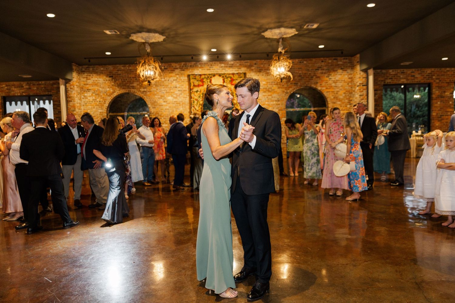 Couple shares first dance in rustic stone reception venue with chandeliers and guests in background.