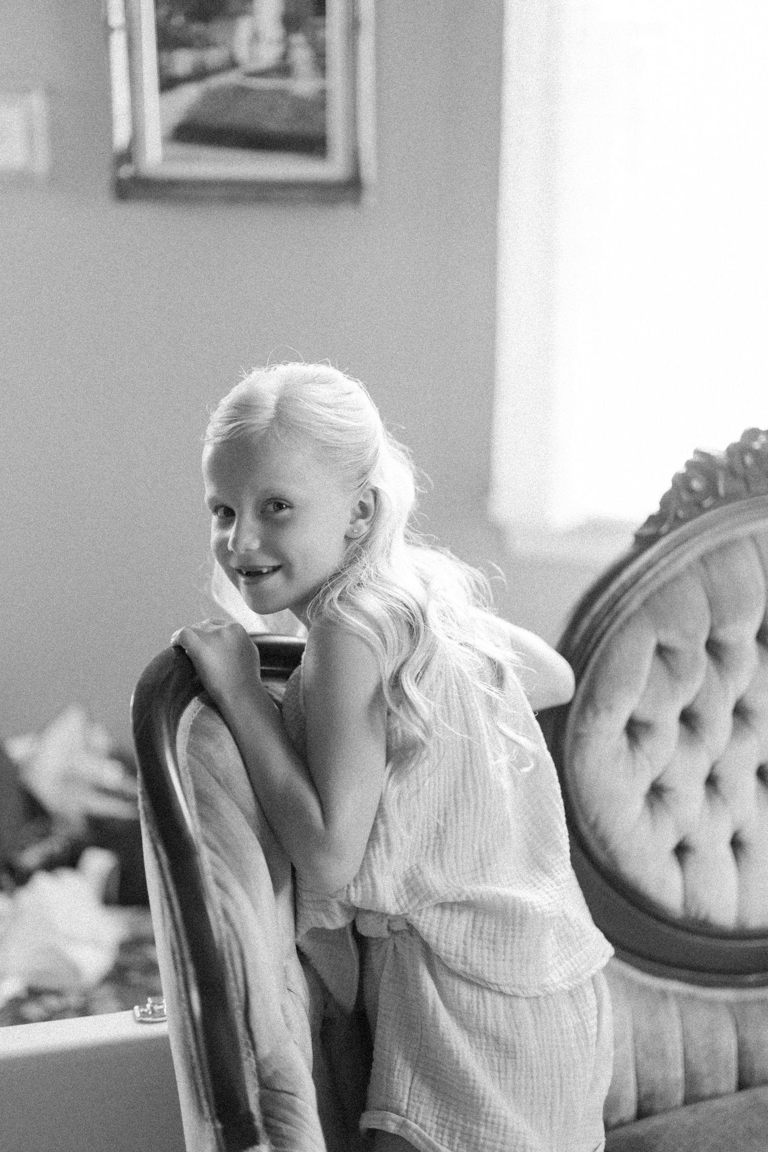 Black and white photo of a smiling young blonde girl in a white dress leaning on a vintage tufted chair indoors.