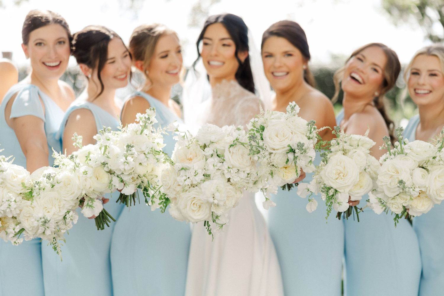 Bridesmaids in light blue dresses holding white floral bouquets with bride in white gown outdoors.