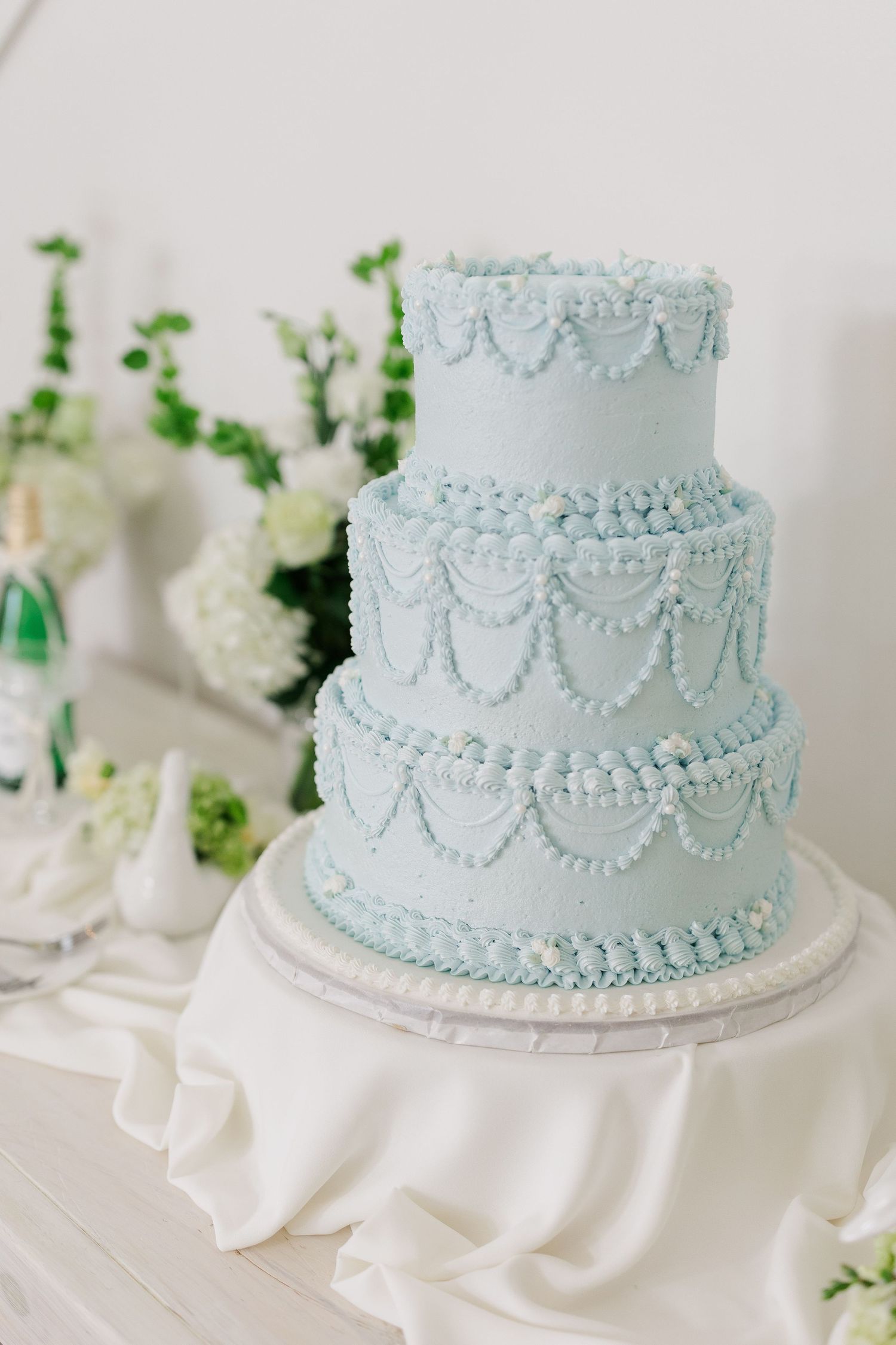 Elegant three-tier light blue wedding cake with pearl swag piping details on a white draped table.
