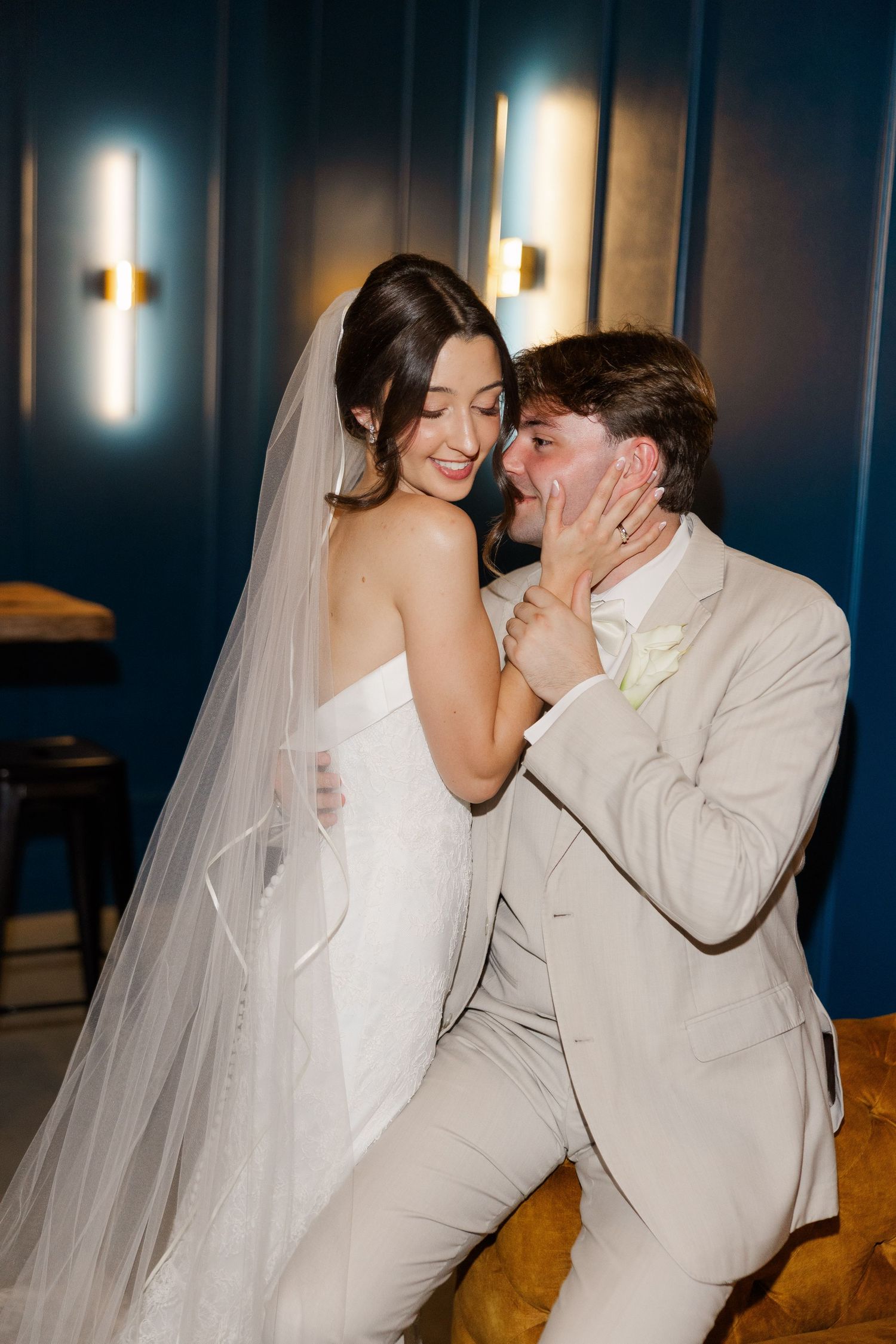 Playful bride in white veil touching grooms face as they laugh together in blue-lit reception room.