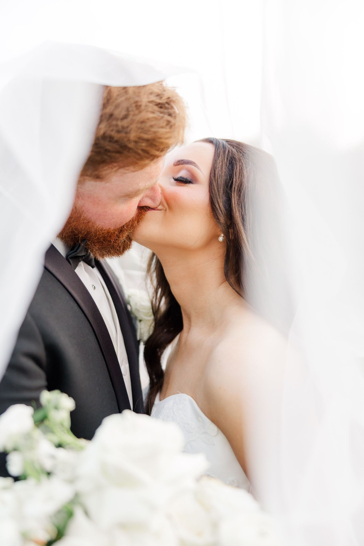 Romantic close-up of couple kissing under wedding veil with white flowers in foreground.