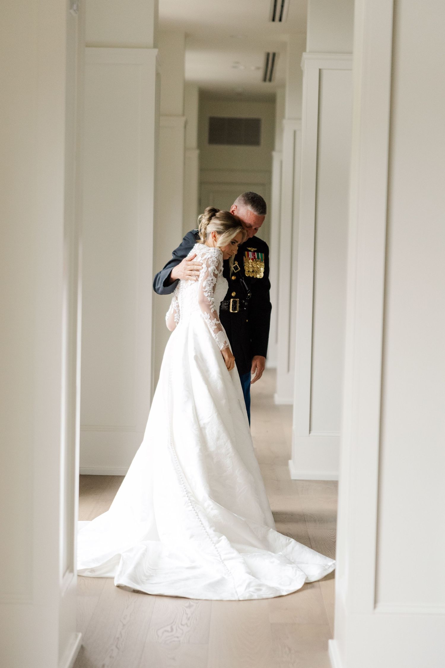 A military service member and bride share an intimate moment in a white hallway on their wedding day.