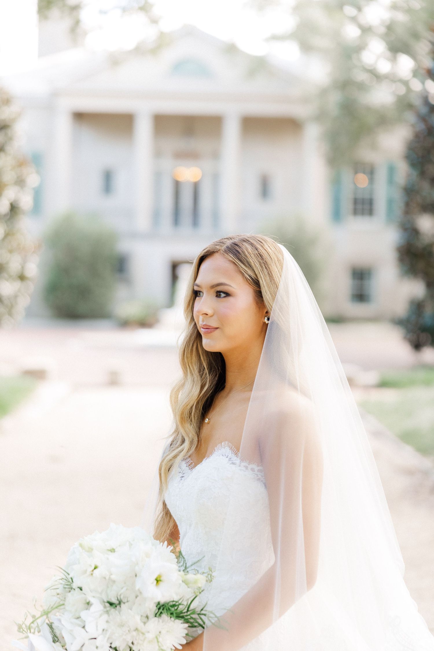A bride wearing a veil and strapless white gown holds a white floral bouquet in front of a colonial-style house.