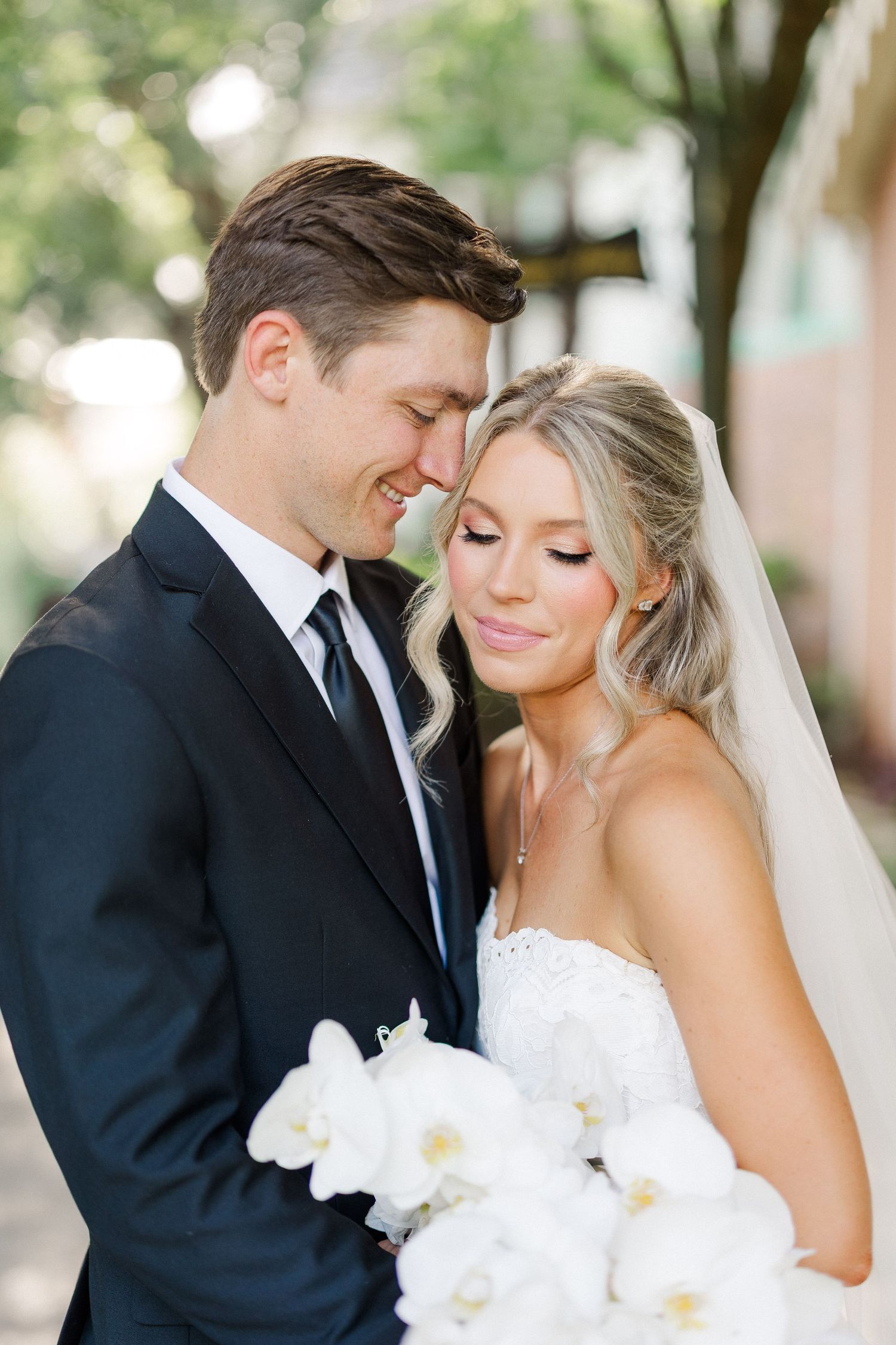 Romantic couple portrait of bride and groom touching foreheads while bride holds white orchid bouquet outdoors.