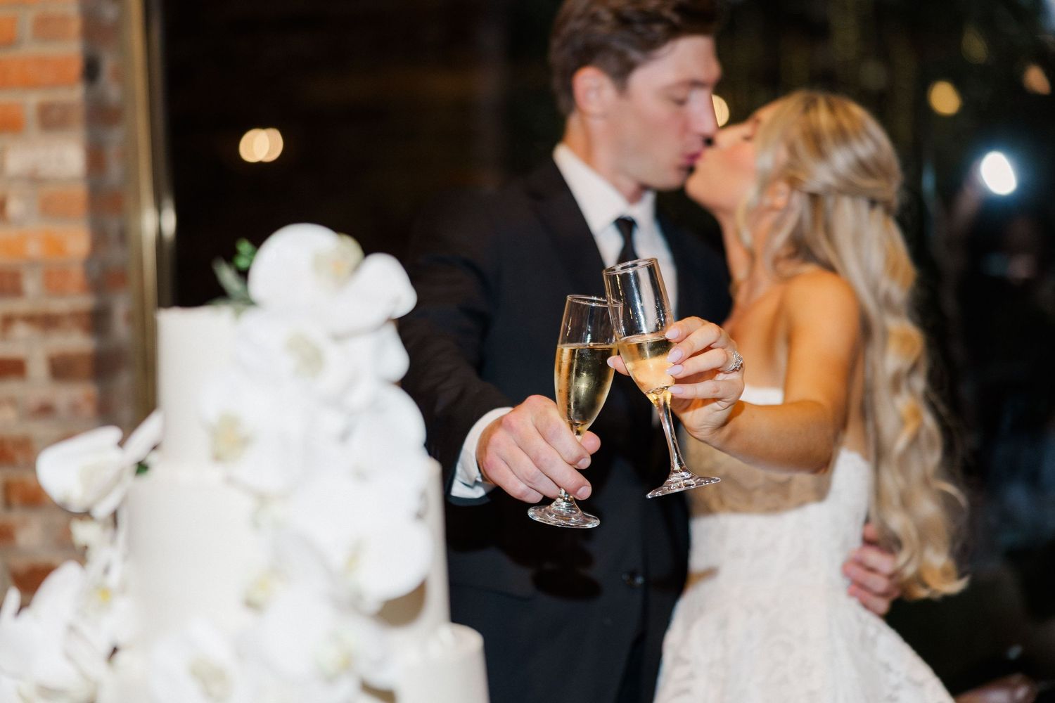 Newlyweds kiss while clinking champagne glasses beside a white orchid-adorned wedding cake.