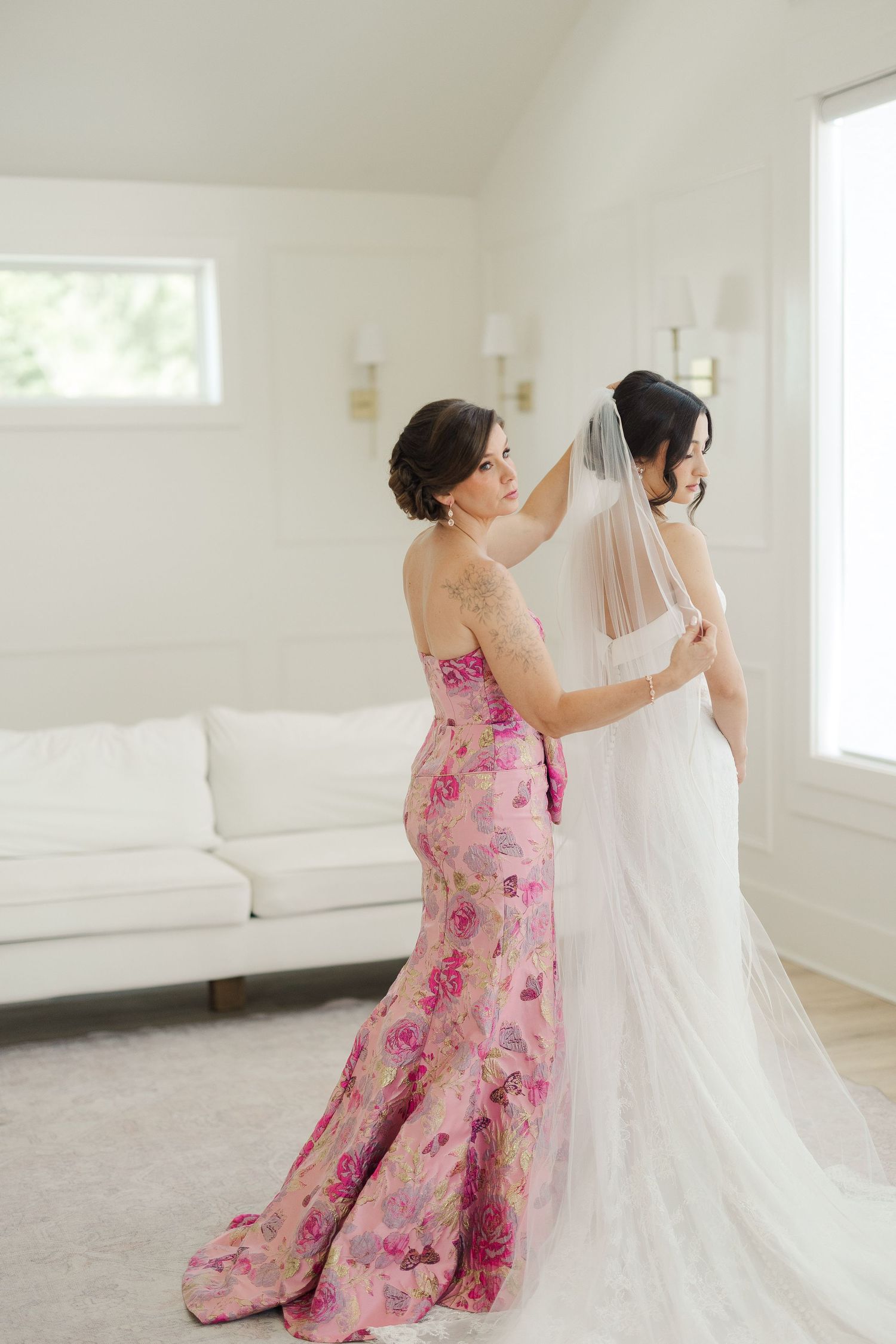 Bridesmaid in pink floral gown adjusting bride's cathedral veil in bright white bridal suite.