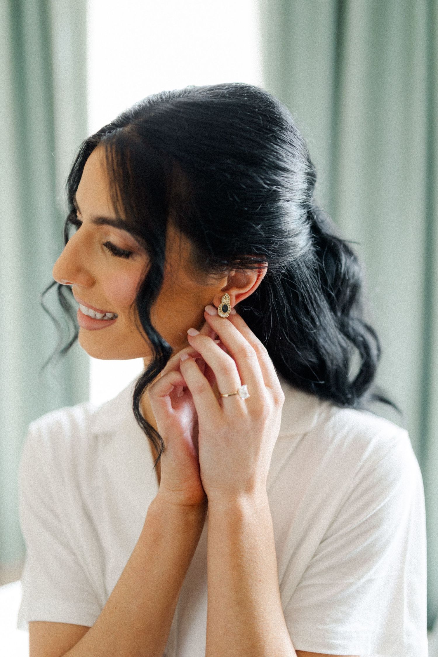Smiling dark-haired bride putting on earrings in white robe before wedding ceremony.