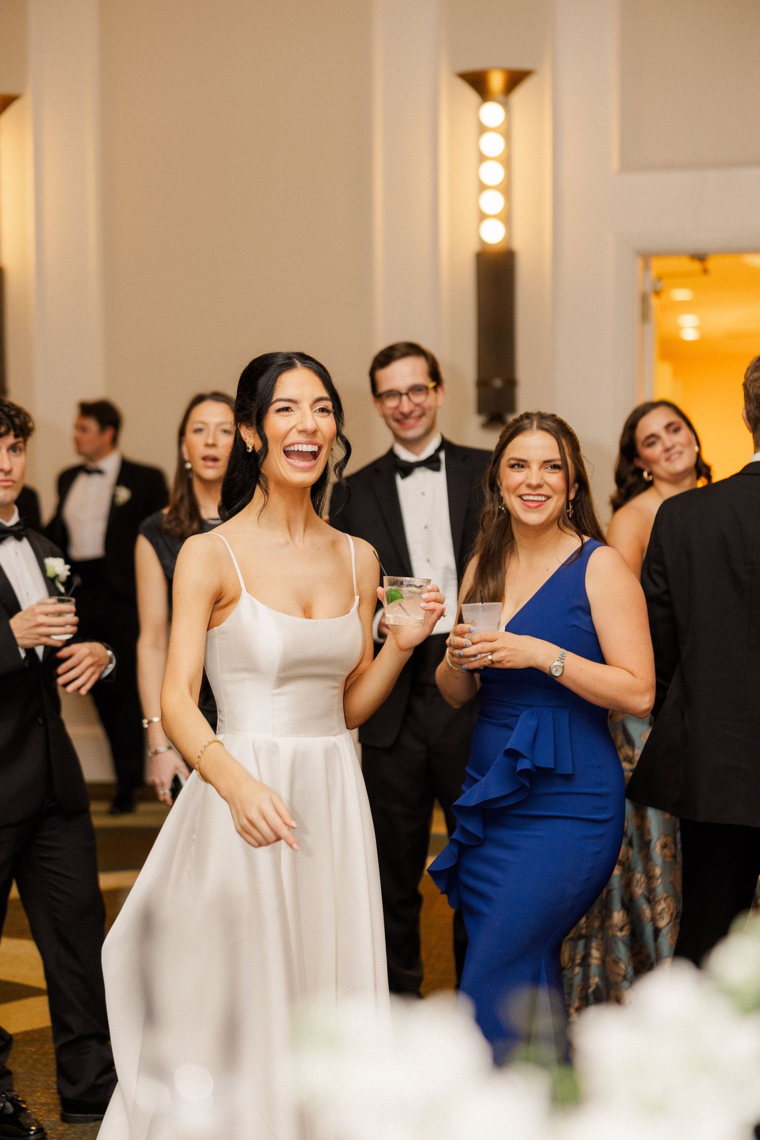 Laughing bride in white gown celebrating with guests in formal attire at wedding reception.