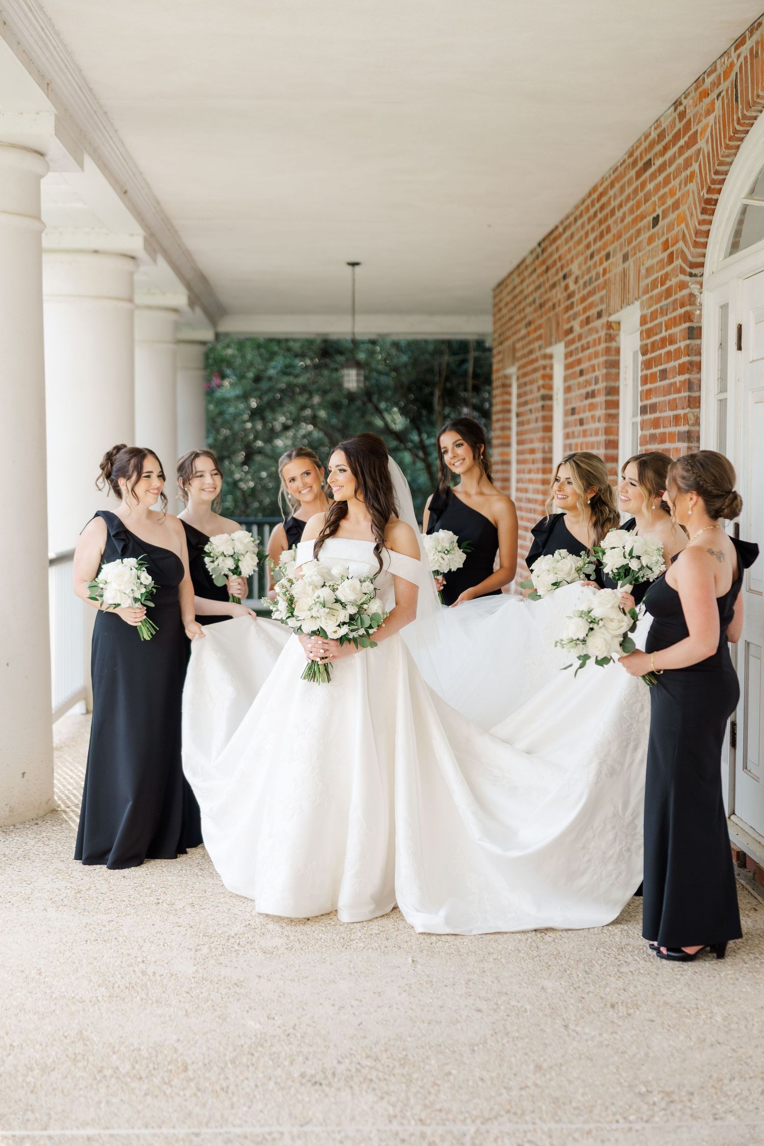 Bridesmaids in black dresses hold up the bride's wedding gown train in a covered outdoor walkway.