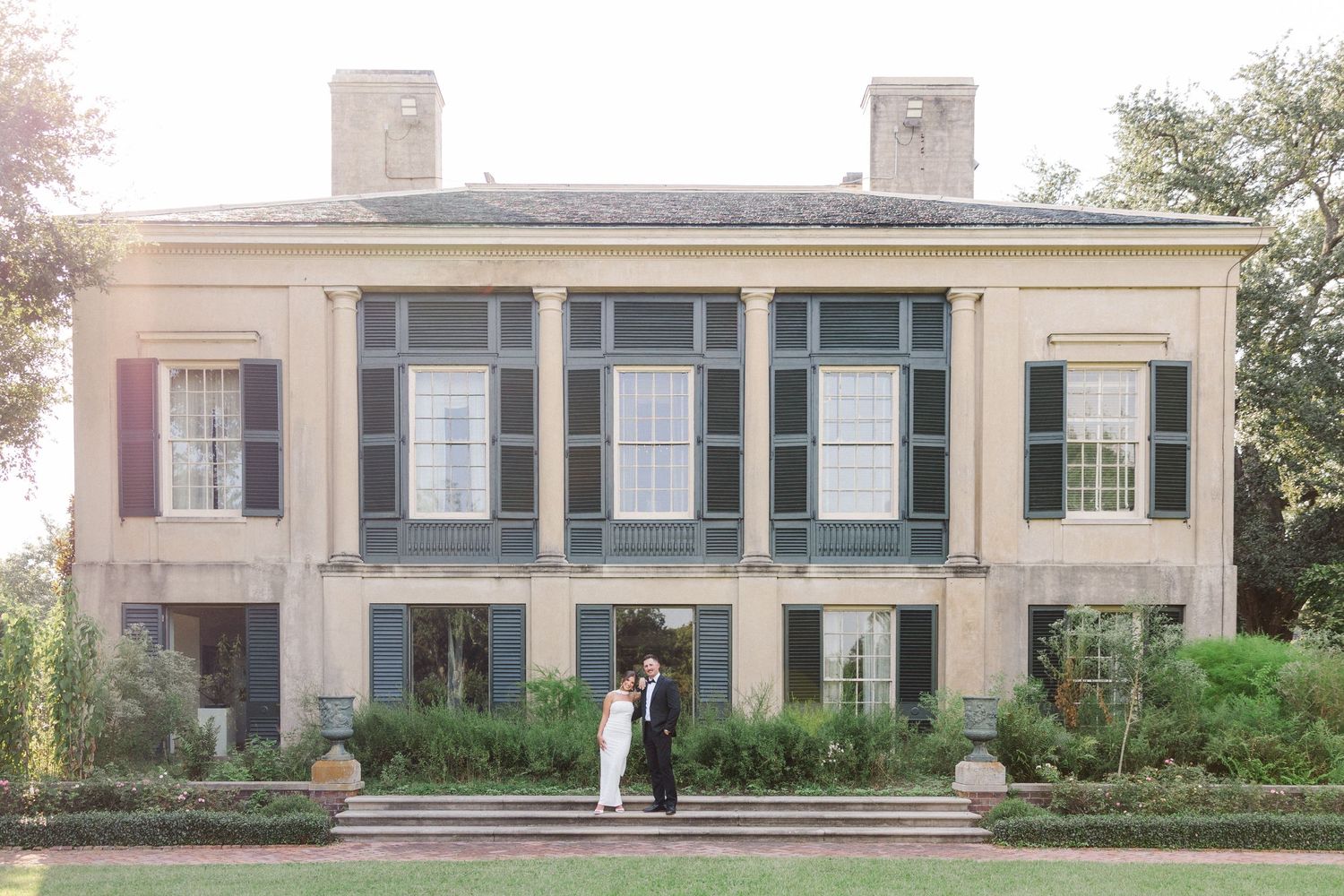 Elegant cream-colored building with white columns and large windows set against a landscaped garden on a sunny day.