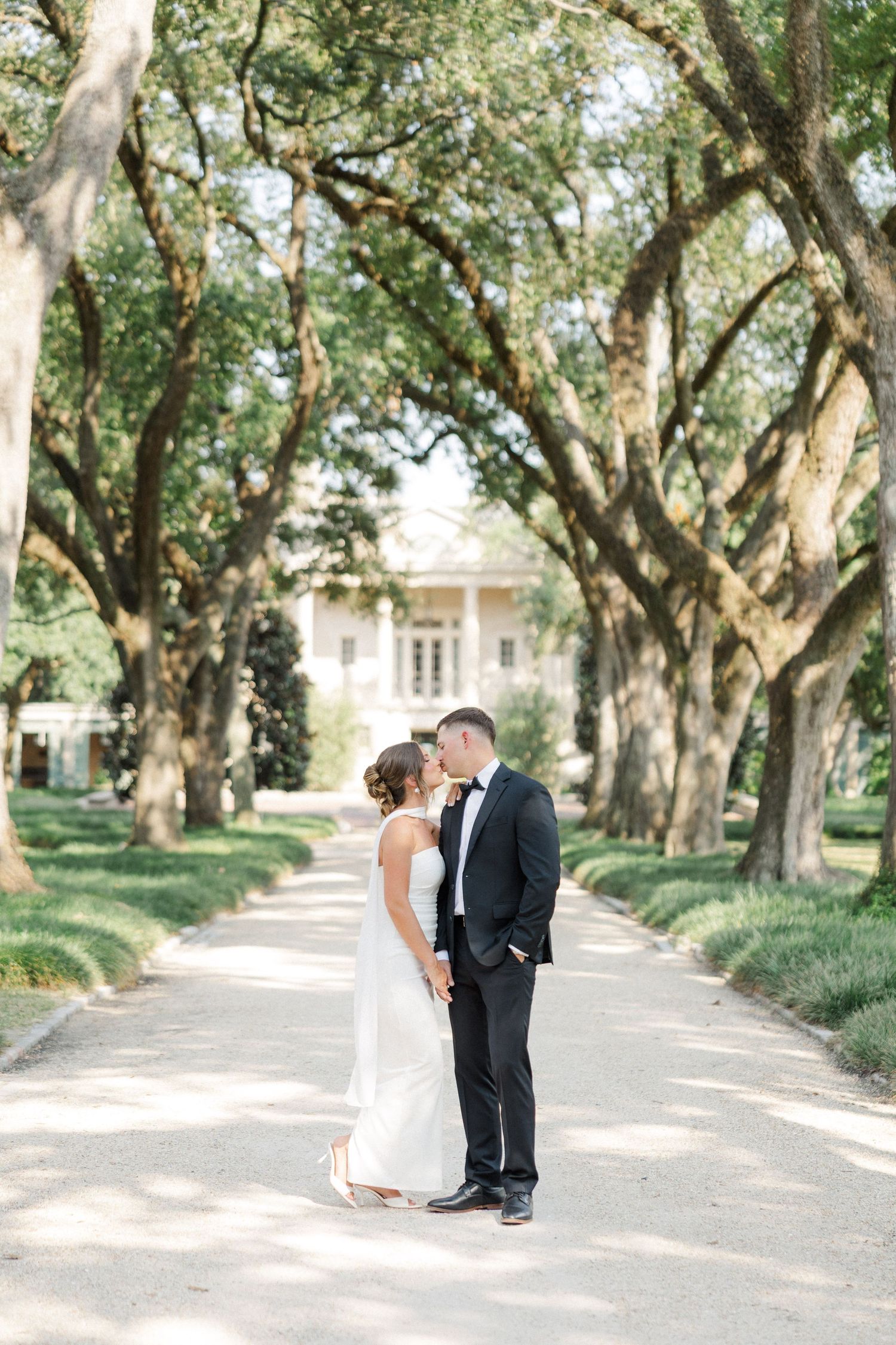 A romantic wedding couple stands on a tree-lined path leading to a stately white mansion on a sunny day.