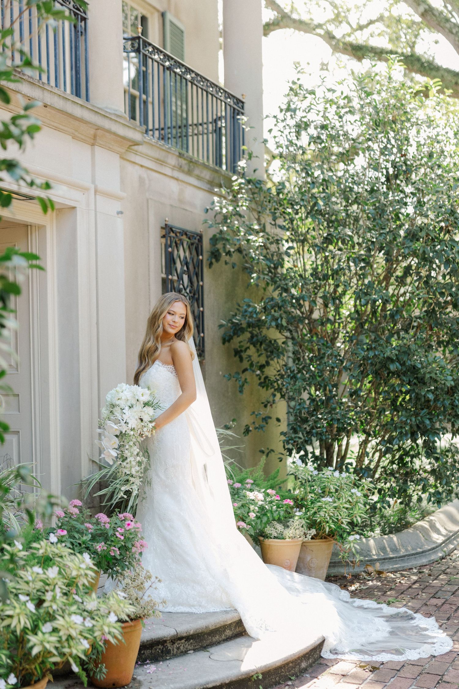 Elegant wedding photo on garden steps with flowing white dress and lush greenery surrounding mansion exterior.